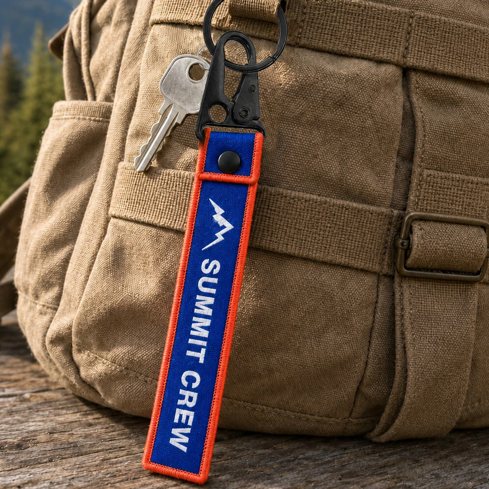 Custom woven Summit Crew keychain clipped to a backpack outdoors